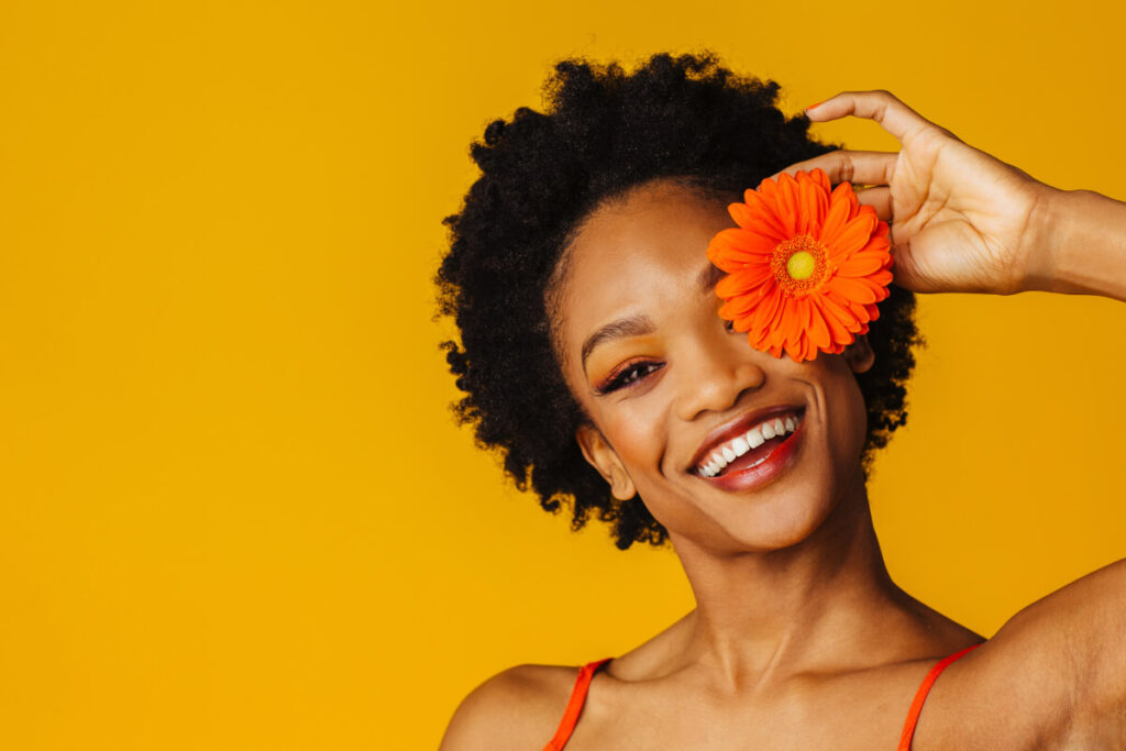 Portrait of a happy excited young woman holding orange gerbera daisy covering her eye and smiling at camera