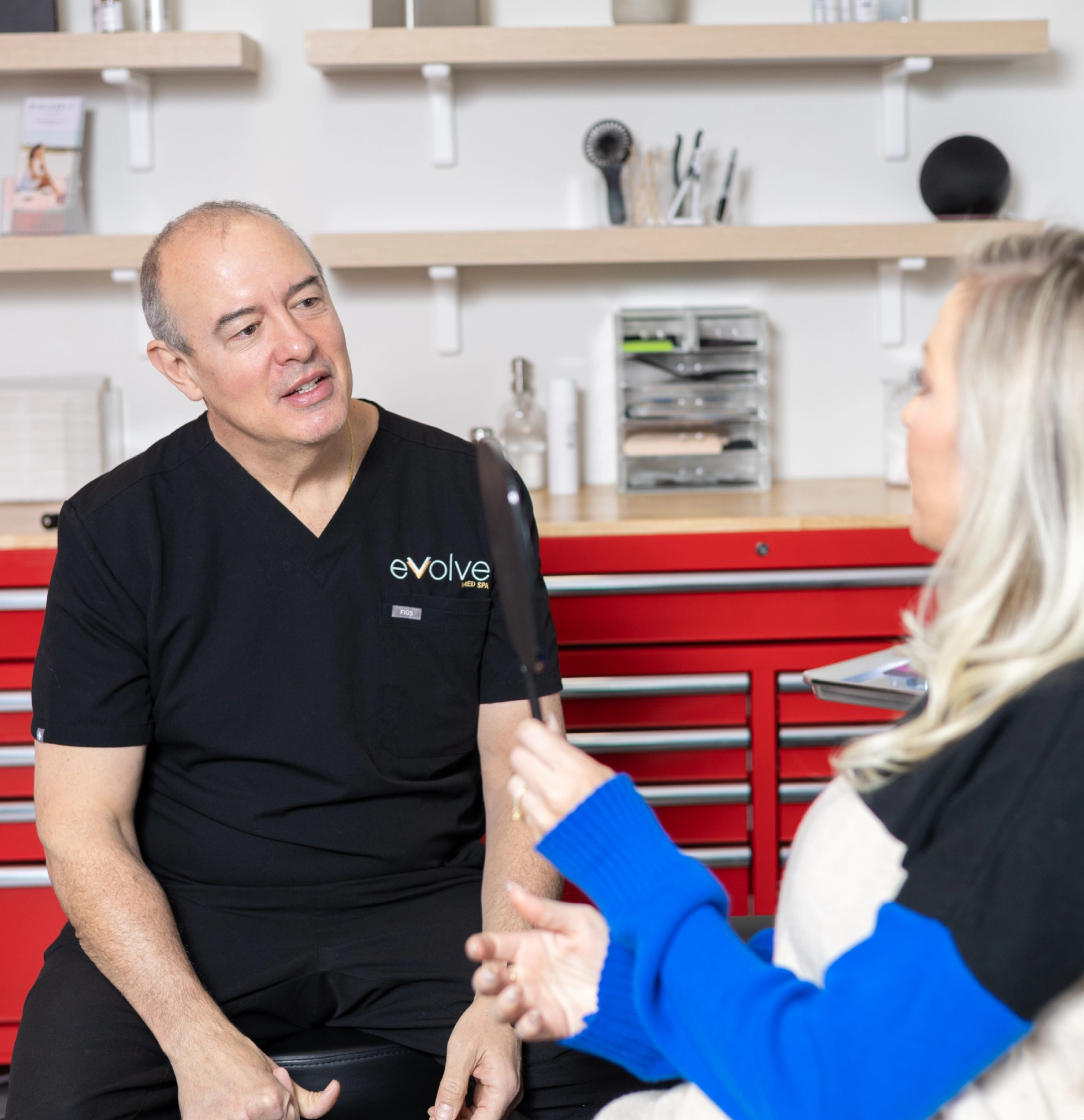 A male Evolve technician in black scrubs with an Evolve logo listens to a client holding a handheld mirror.