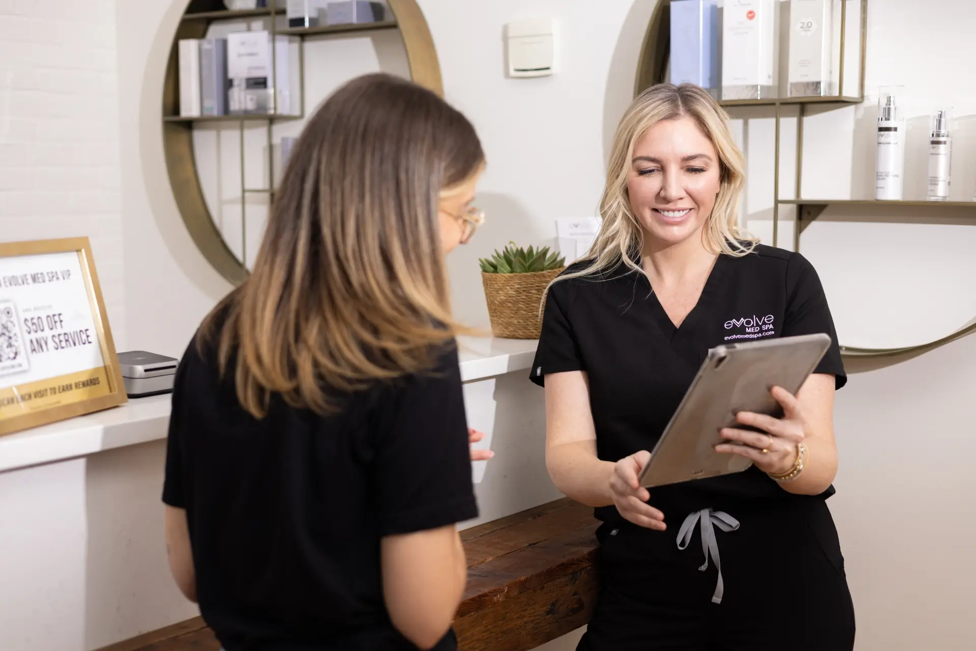 A Evolve professional wearing a black uniform showing a tablet to a client at a reception area, with shelves of skincare products and a promotional sign visible in the background, creating a warm and professional atmosphere.