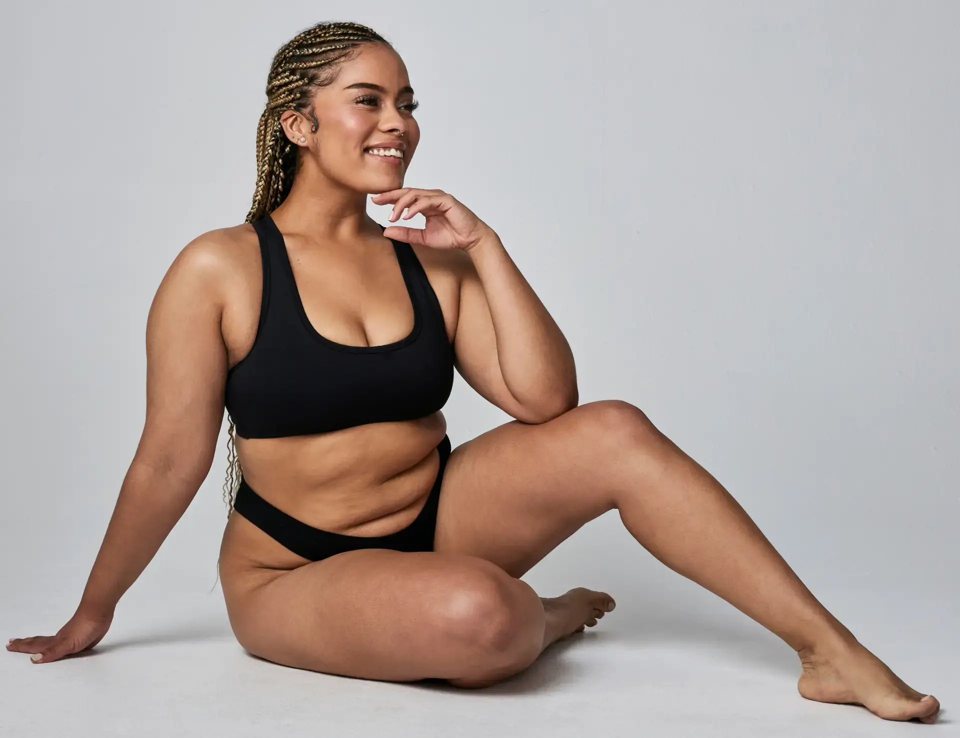 A confident woman with braided hair sitting on the floor, wearing a black sports bra and briefs, smiling and posing.