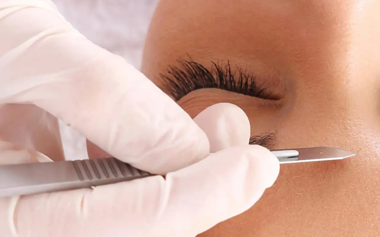 Close-up of a dermaplaning treatment being performed, with a gloved hand holding a sterile scalpel gently exfoliating the skin of a person with closed eyes and long eyelashes.