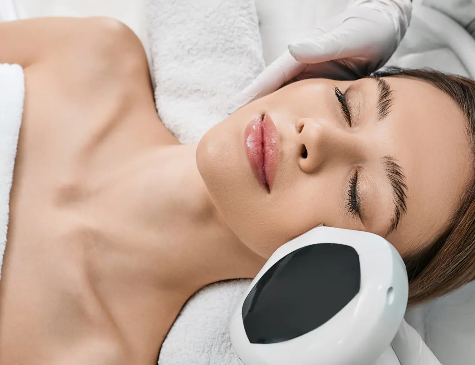 Woman receiving a relaxing LED light facial treatment, lying down with closed eyes as a device gently touches her cheek, promoting skin rejuvenation.