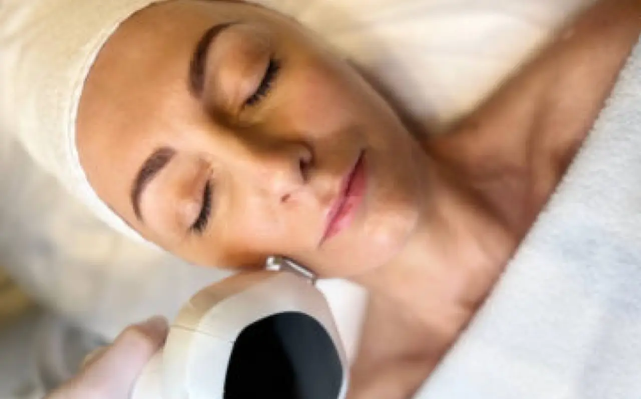 A person undergoing an IPL (Intense Pulsed Light) treatment, with their eyes closed, wearing a white headband, and a handheld IPL device applied to their cheek for skin rejuvenation.