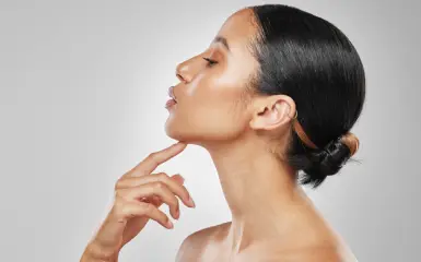 A woman with smooth, glowing skin tilting her head upward and touching her chin, showcasing a defined jawline and confidence.