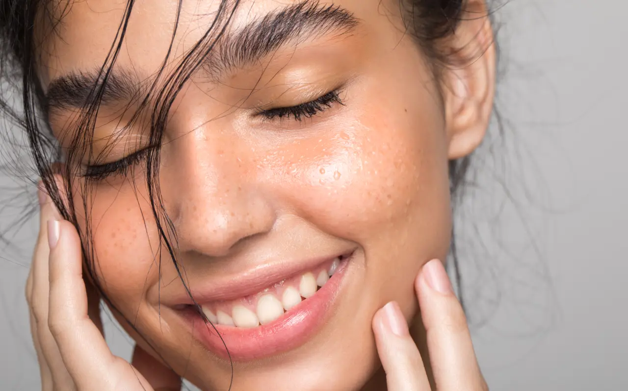 Close-up of a smiling person with glowing, hydrated skin, soft freckles, and slightly damp hair framing their face, showcasing a fresh and radiant complexion.