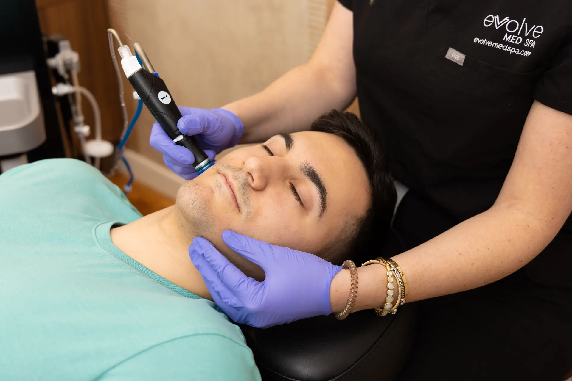 A man receiving a HydraFacial treatment at a med spa, lying on a chair with his eyes closed, while a professional wearing purple gloves uses a specialized device on his chin for deep cleansing and hydration.